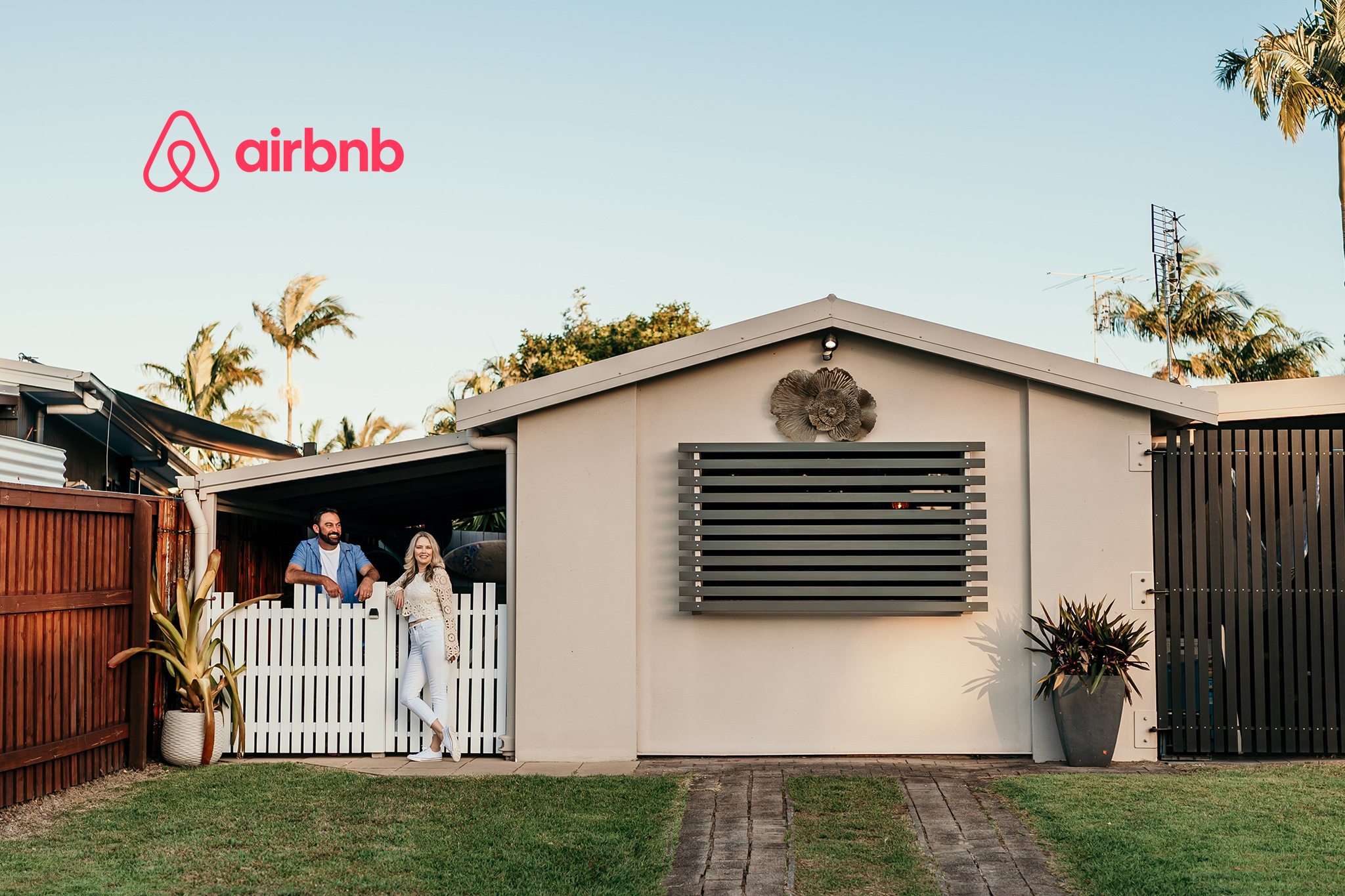 A small modern guest house with a white gate, two people standing in the yard, and an Airbnb logo in the upper left corner.