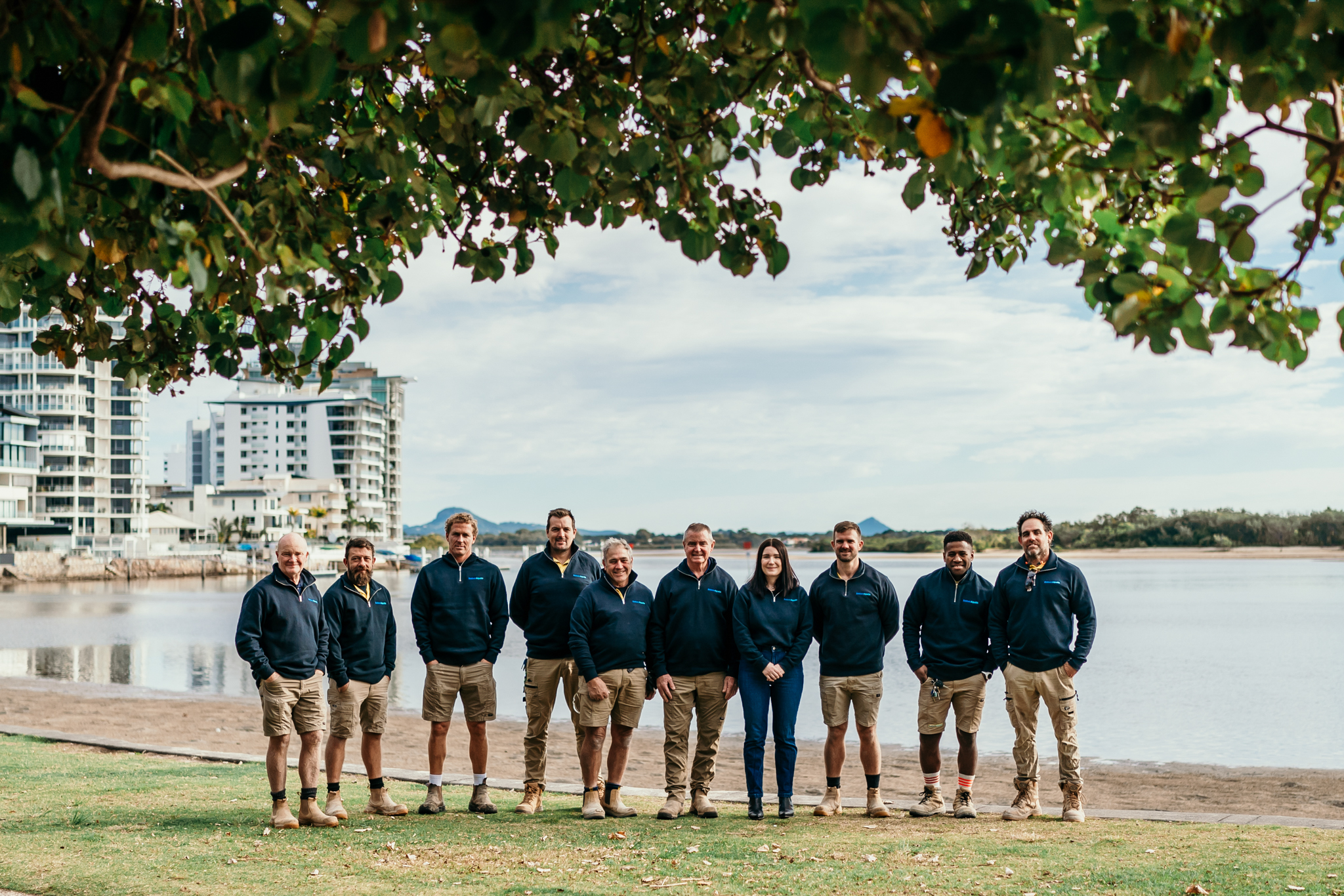 Group of people in navy hoodies and khaki shorts stand in a line on a grassy waterfront, with leafy branches overhead and a cityscape in the background.