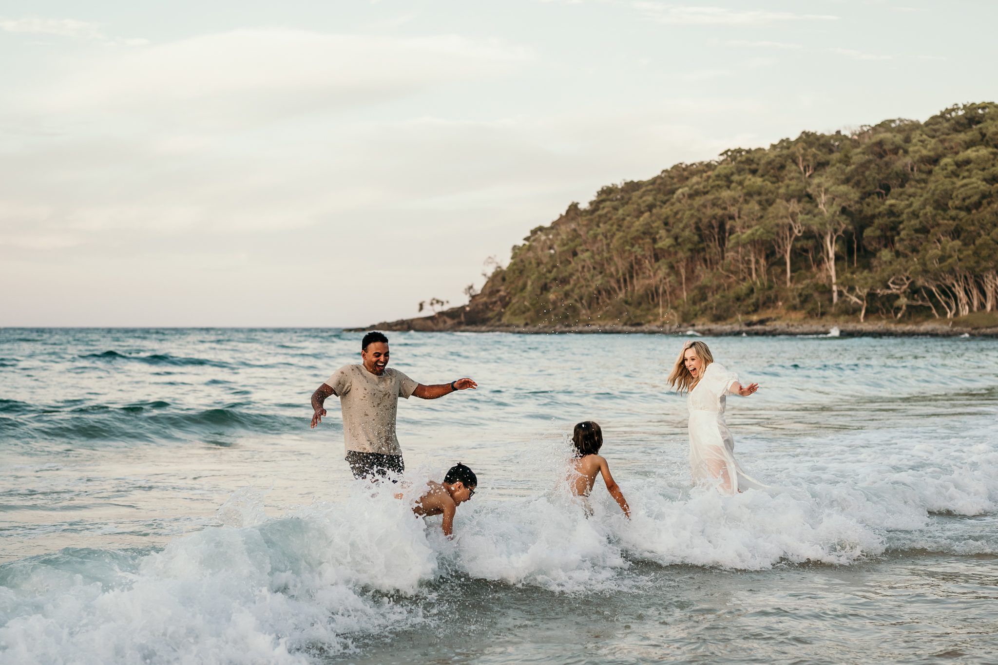 Two adults and two children splash in the shallow waves at the beach, with a forested hill in the background.
