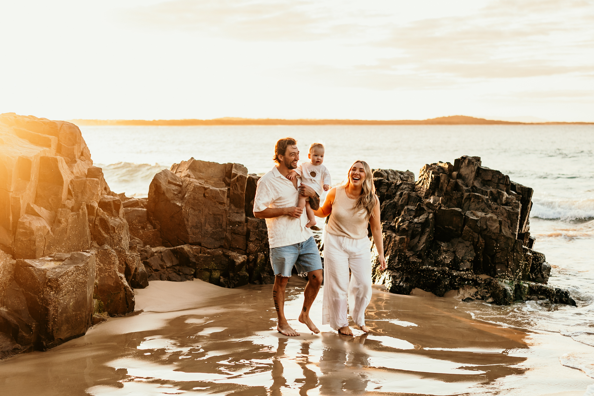 Three people walk along a rocky beach at sunset with the ocean in the background.