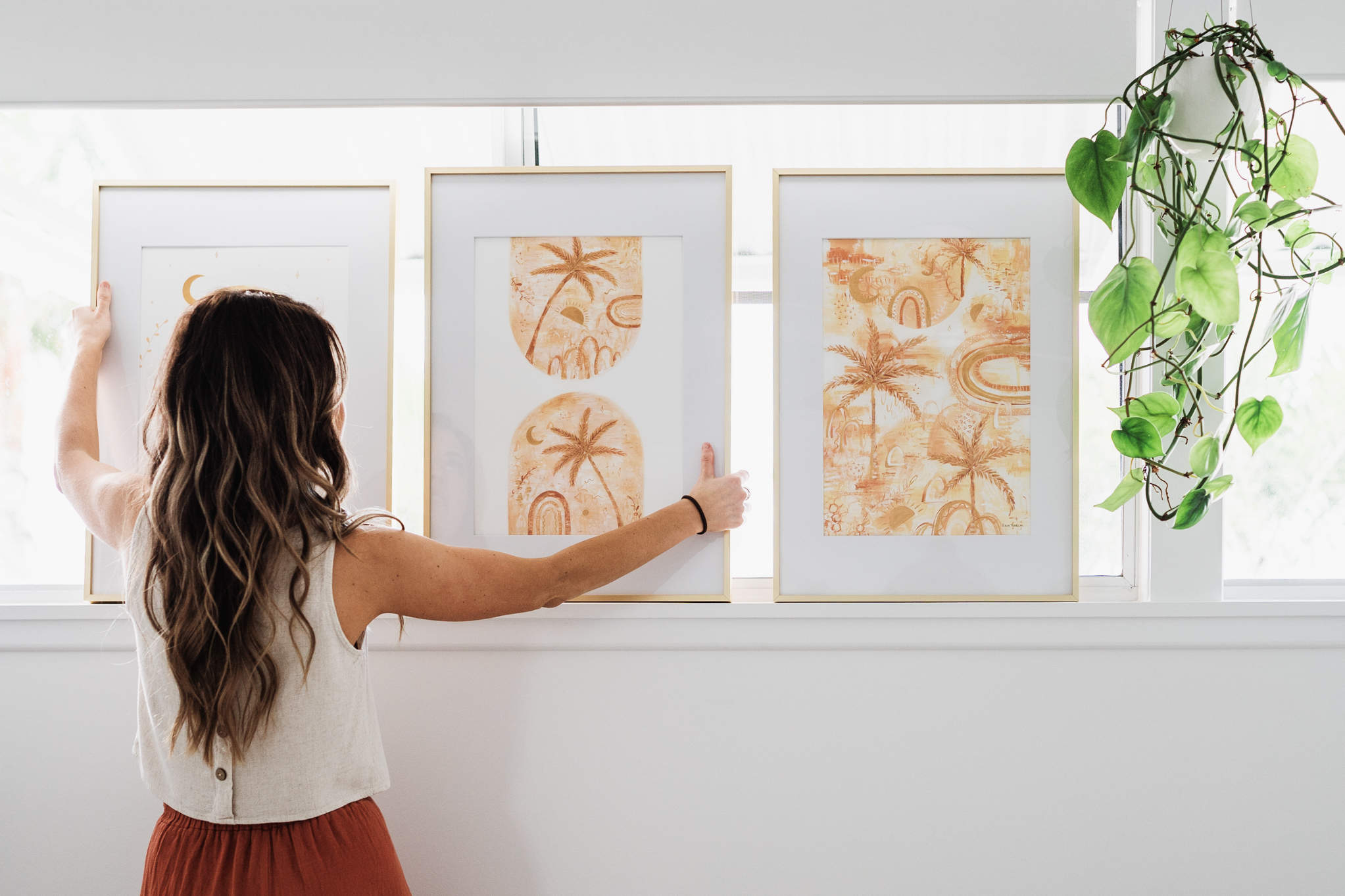 A woman with long hair faces away, adjusting three framed orange botanical prints on a white wall beside a hanging plant.