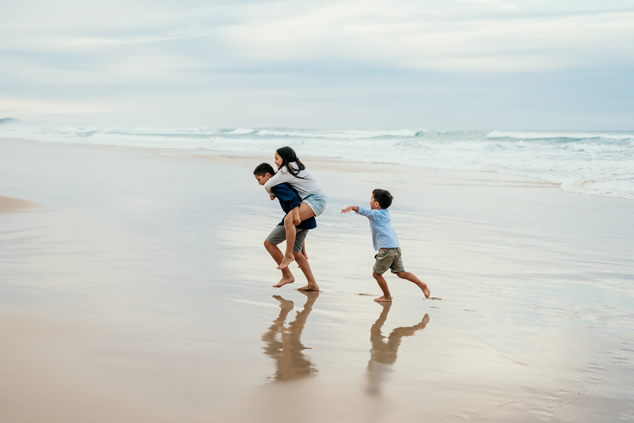 Three people run along the wet sand at the beach with waves in the background.