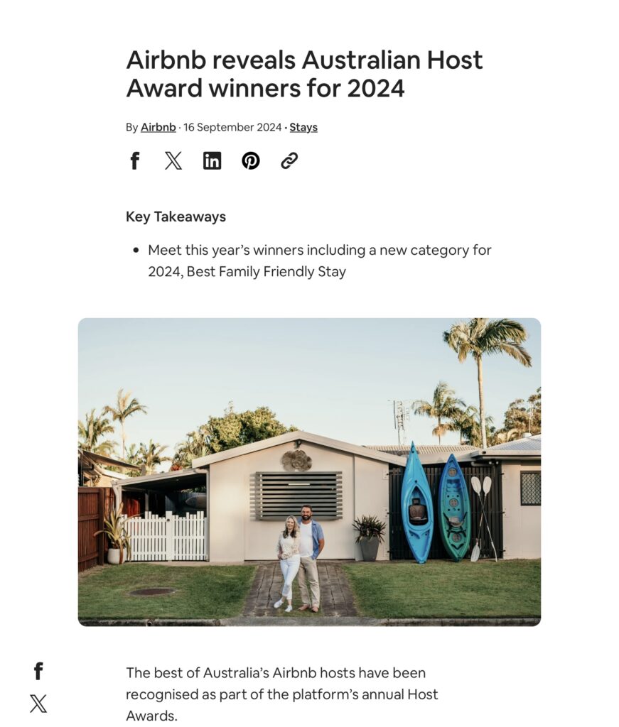 Two people stand in front of a single-story house with two blue surfboards leaned against the wall and palm trees in the yard.