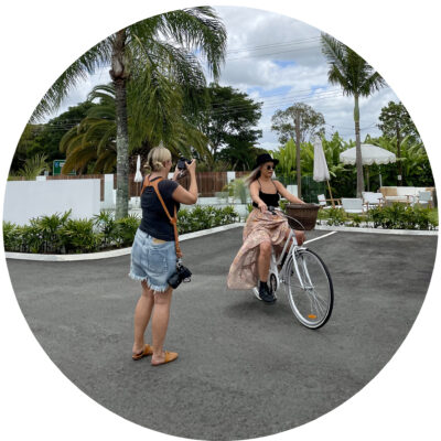 Two women outdoors in a tropical setting with palm trees; one photographs the other as she rides a white bicycle.