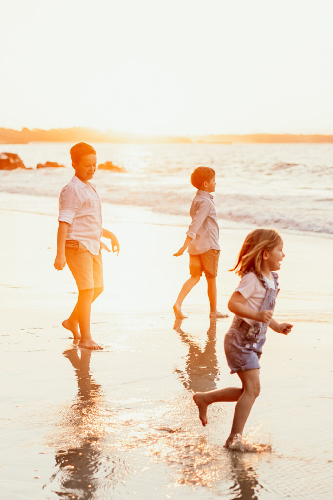 Three children play along a sunlit beach at sunset, with wet sand and gentle waves.