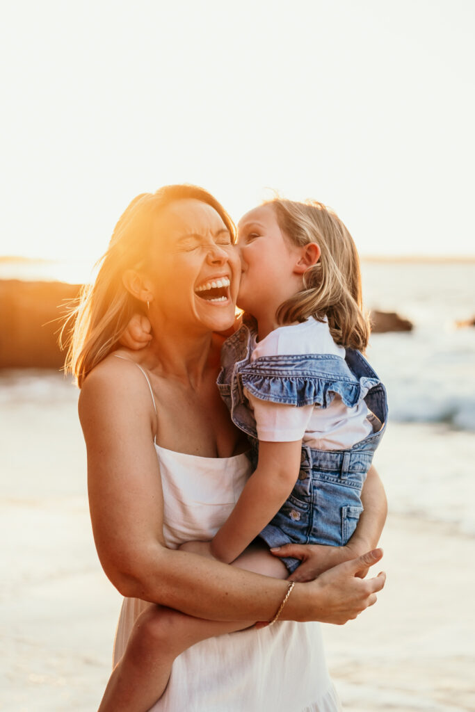 A smiling woman holds a young girl who is kissing her cheek on a sunny beach at sunset.