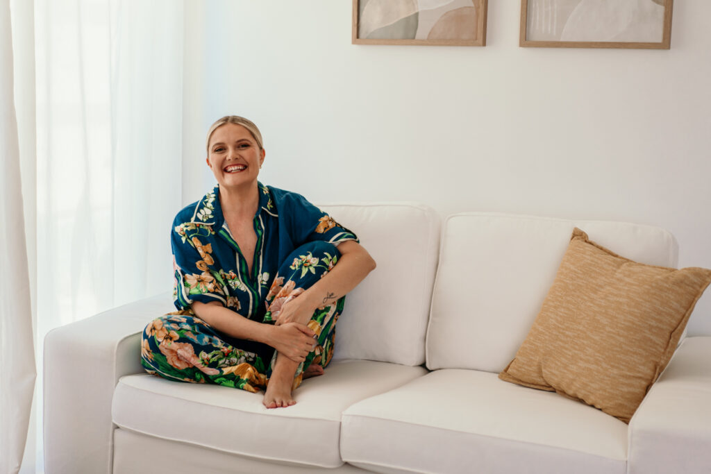 Smiling woman in a blue floral robe sits on a white sofa in a bright living room.
