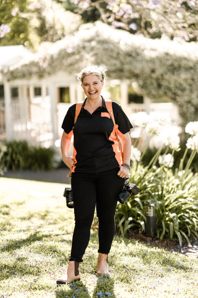 Smiling woman in a black and orange athletic outfit stands on a sunny lawn with a house and greenery in the background.