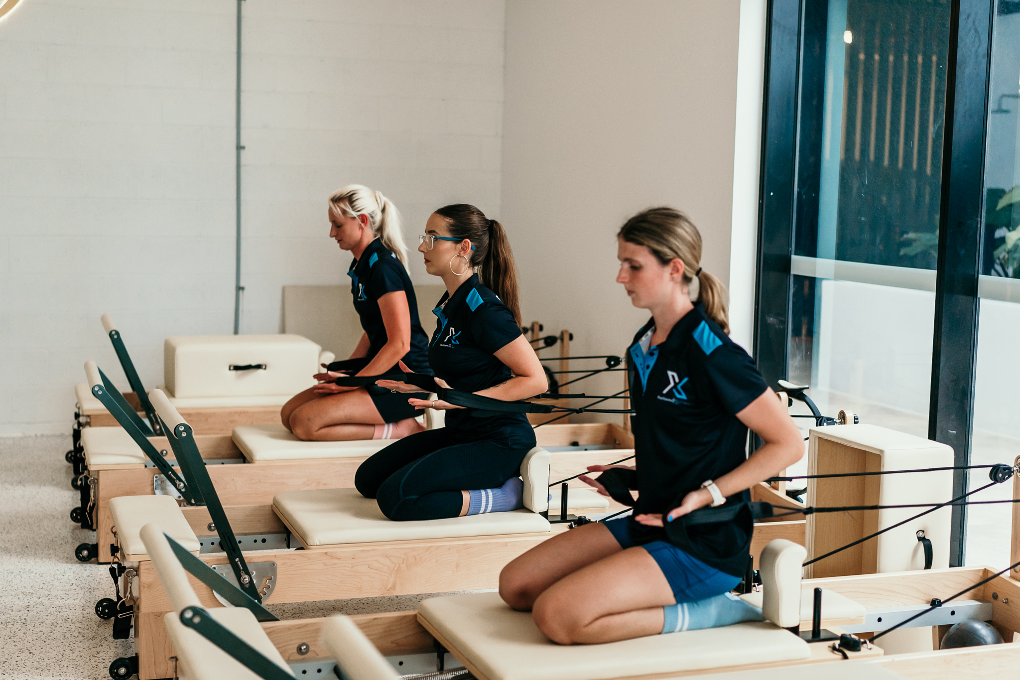 Three women in athletic wear sit on Pilates reformer machines in a bright studio.