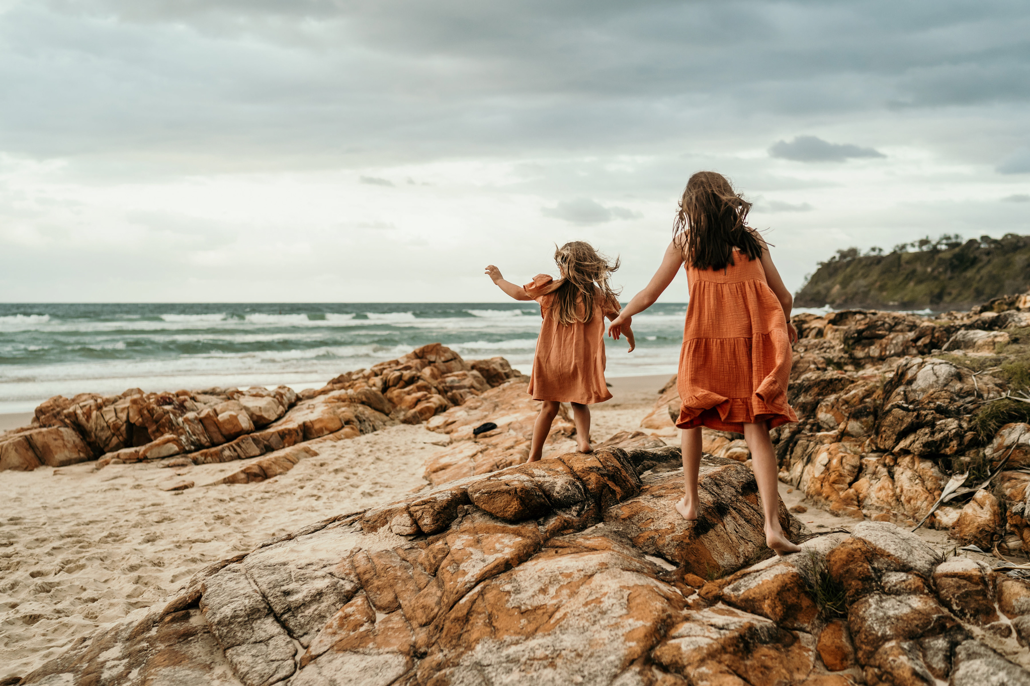 Two girls in orange dresses stand on a rocky beach, holding hands and watching the ocean.