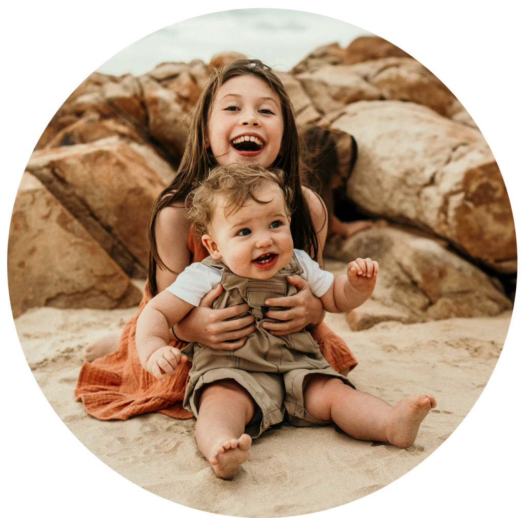 Two smiling children sit on a sandy beach with a rocky background, the older girl holding the baby.