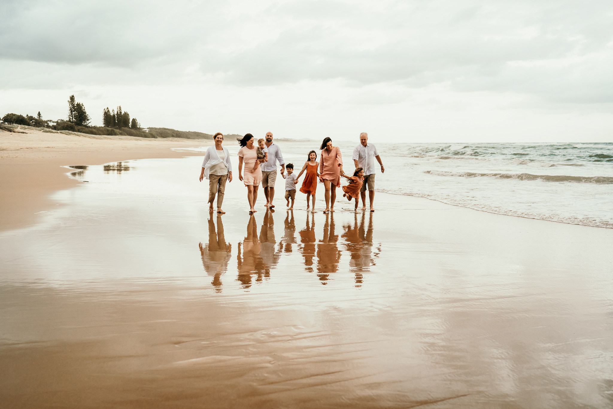 A family group walks along a wet sandy beach by the ocean, with their reflections visible on the shiny surface.