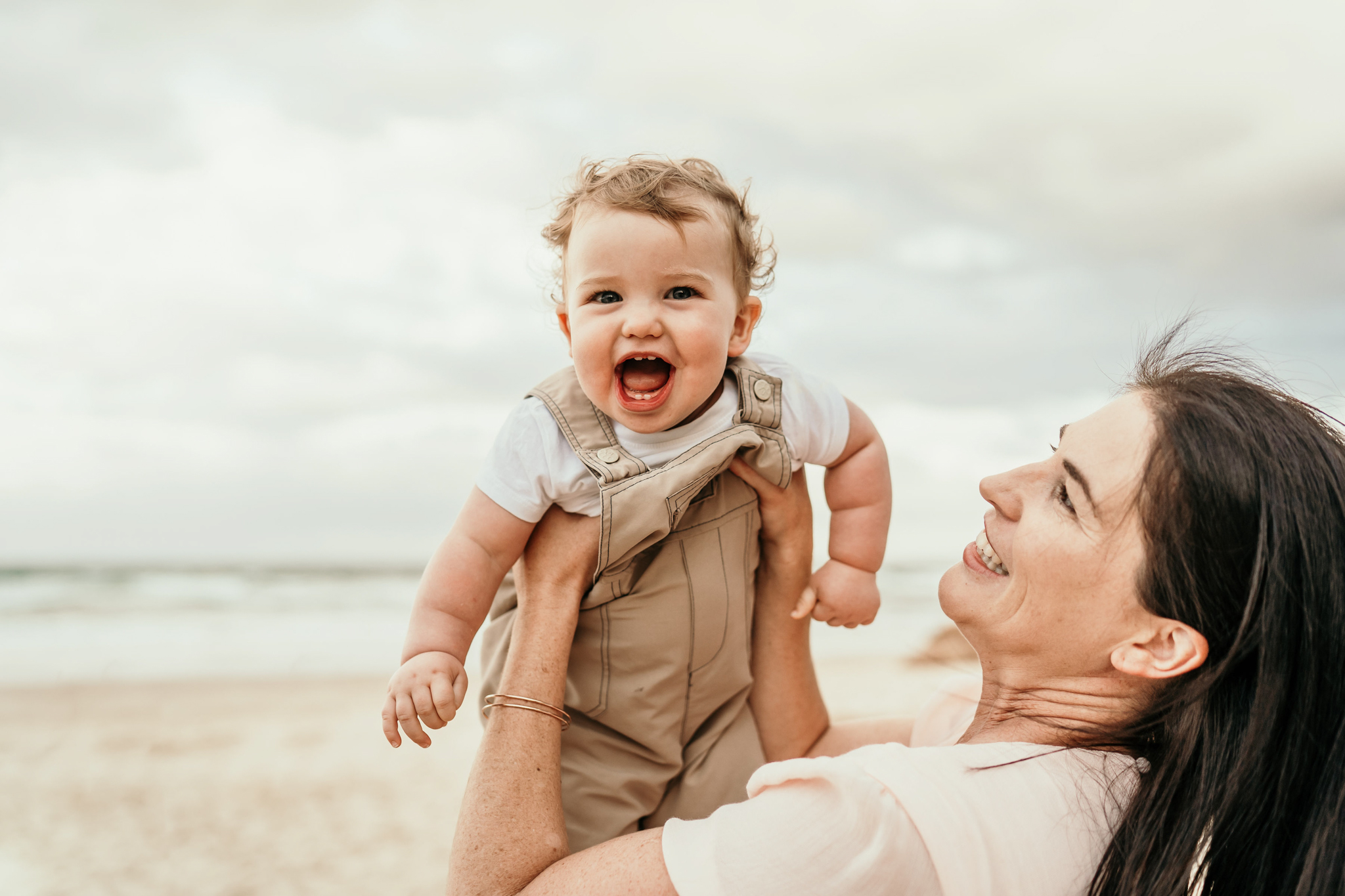 A smiling baby is lifted by a woman on a sandy beach with the ocean in the background.