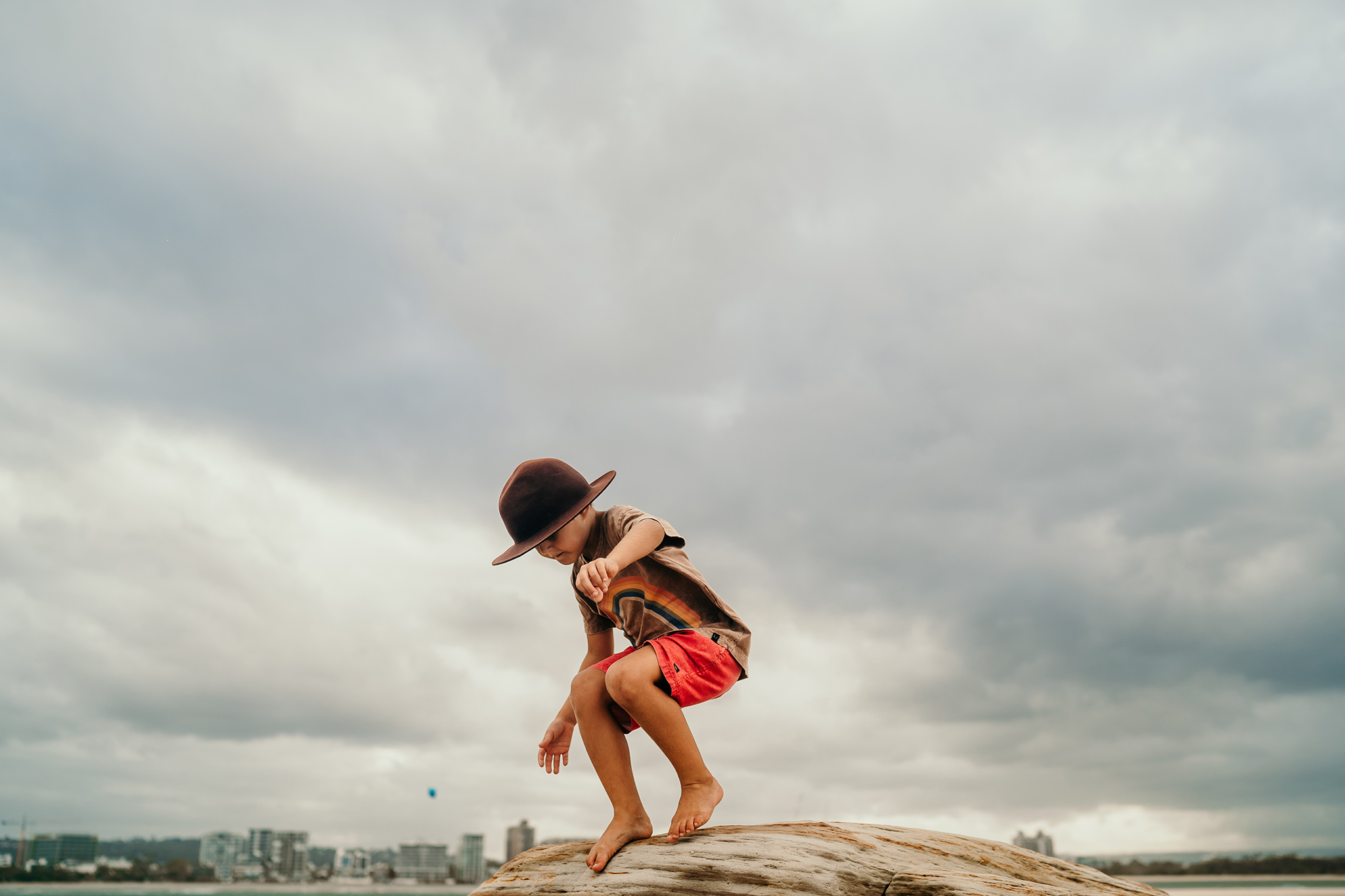 A person wearing a hat and red shorts is jumping on a rocky ledge above the water with a cloudy sky and distant city skyline.