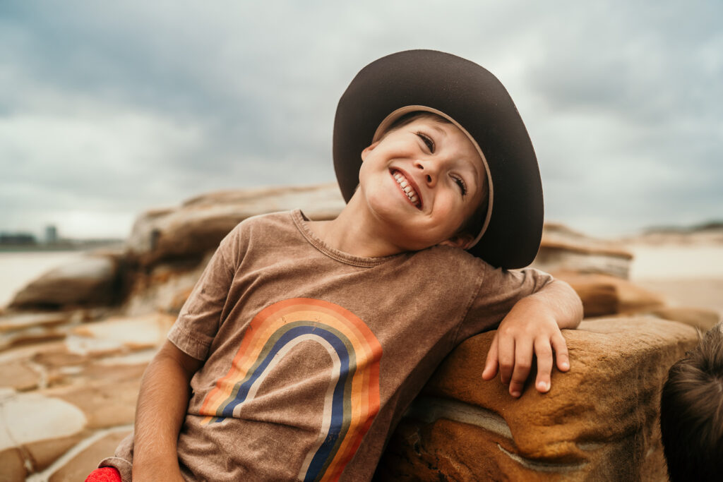 Smiling child wearing a wide-brimmed hat and a T-shirt with a rainbow design leans on a large rock at the beach, with cloudy skies and sandy rocks in the background.