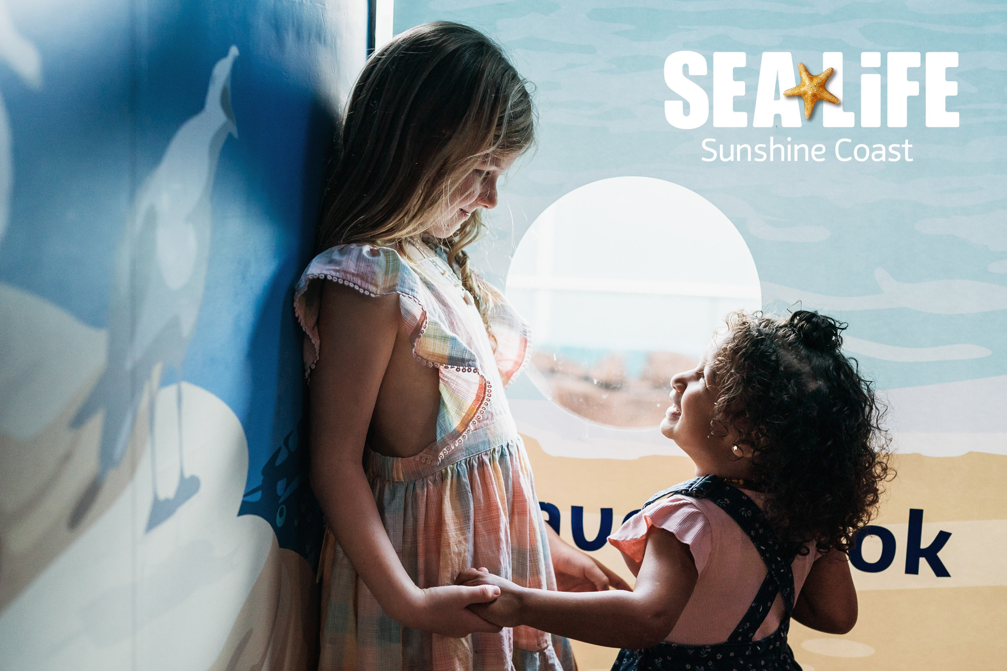 Two young girls stand holding hands and smiling at each other in front of a sunlit SEA LIFE Sunshine Coast aquarium display, with soft light highlighting their faces.