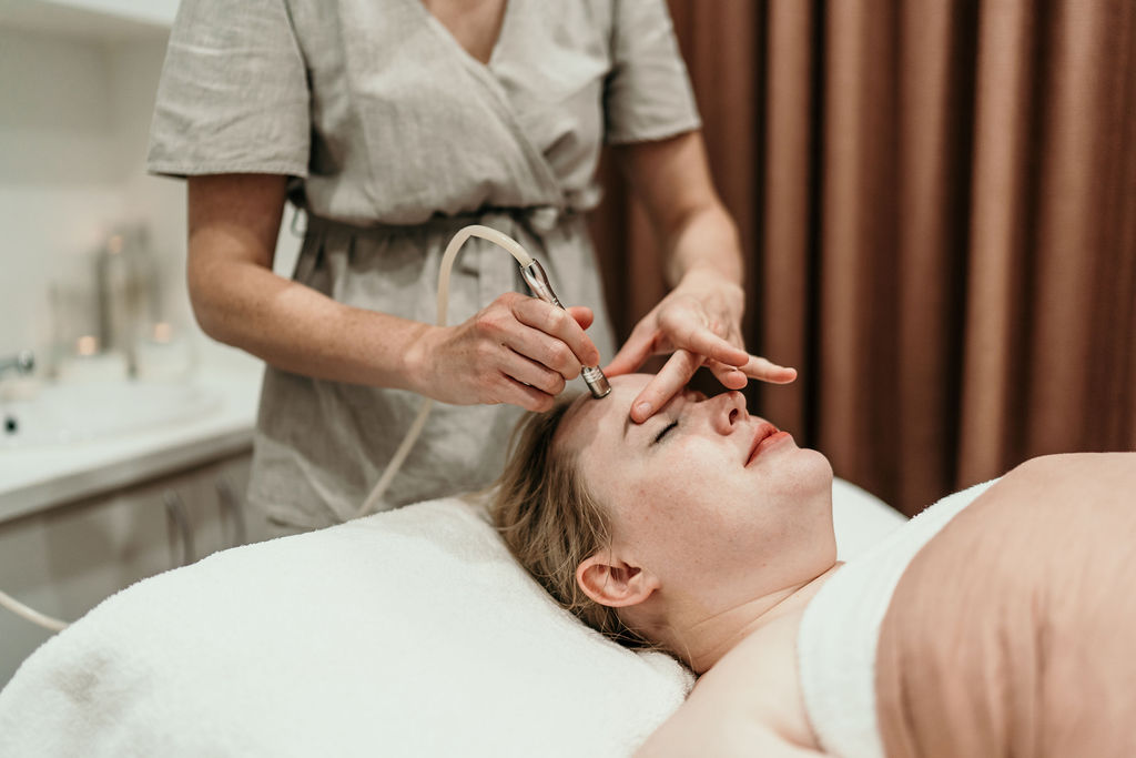 A spa esthetician performs a facial on a client lying on a treatment bed.