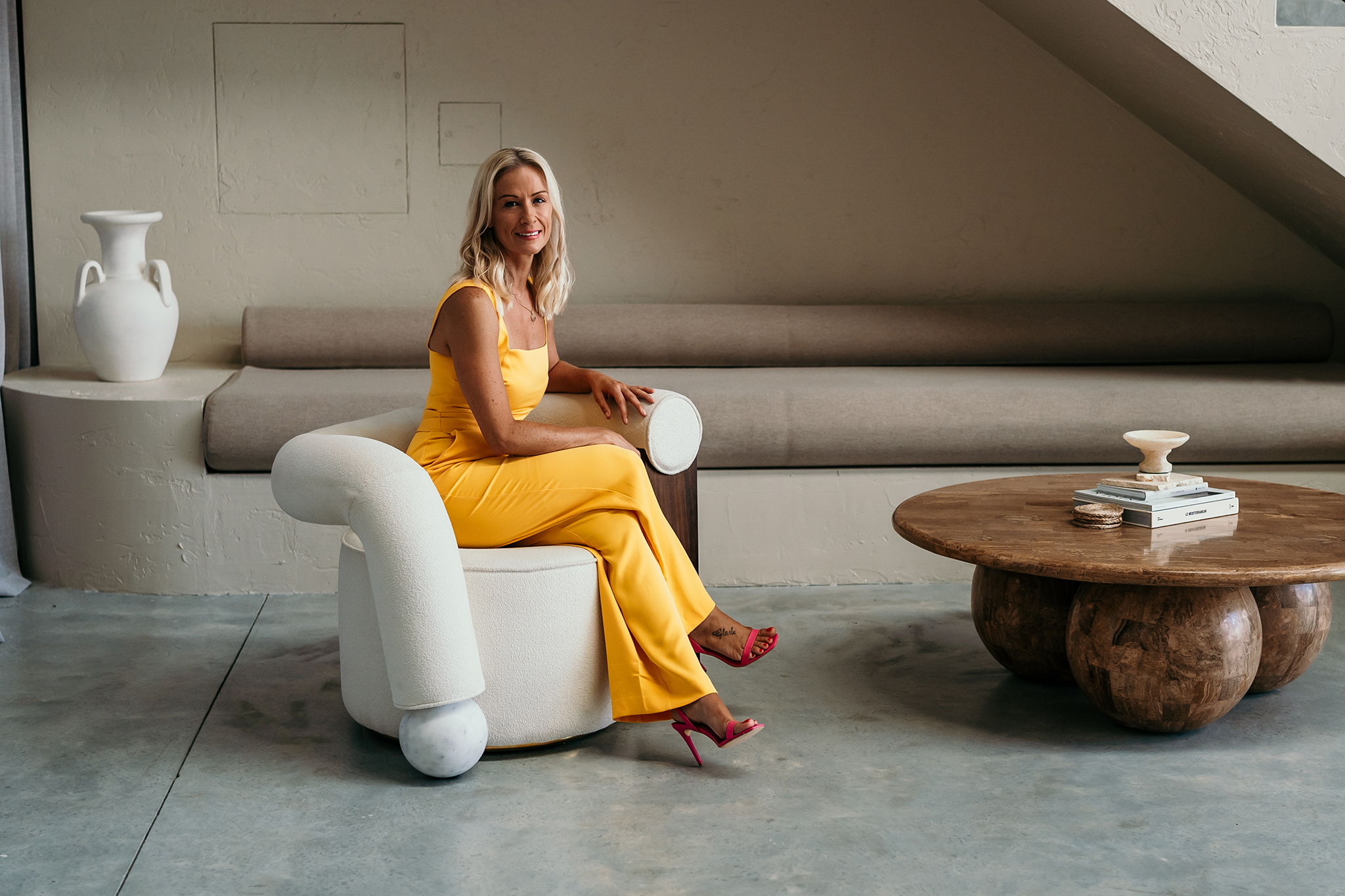 A woman in a yellow outfit sits on a white modern chair in a minimalist living room with a round wooden coffee table.