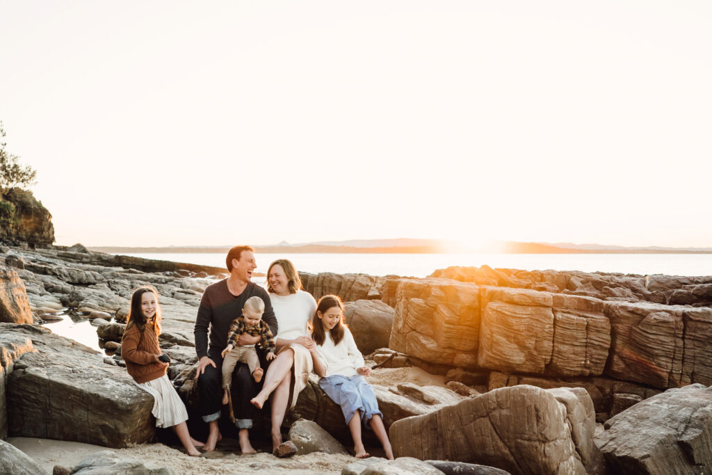 A family sits on large rocks along the shoreline as the sun sets over the ocean.