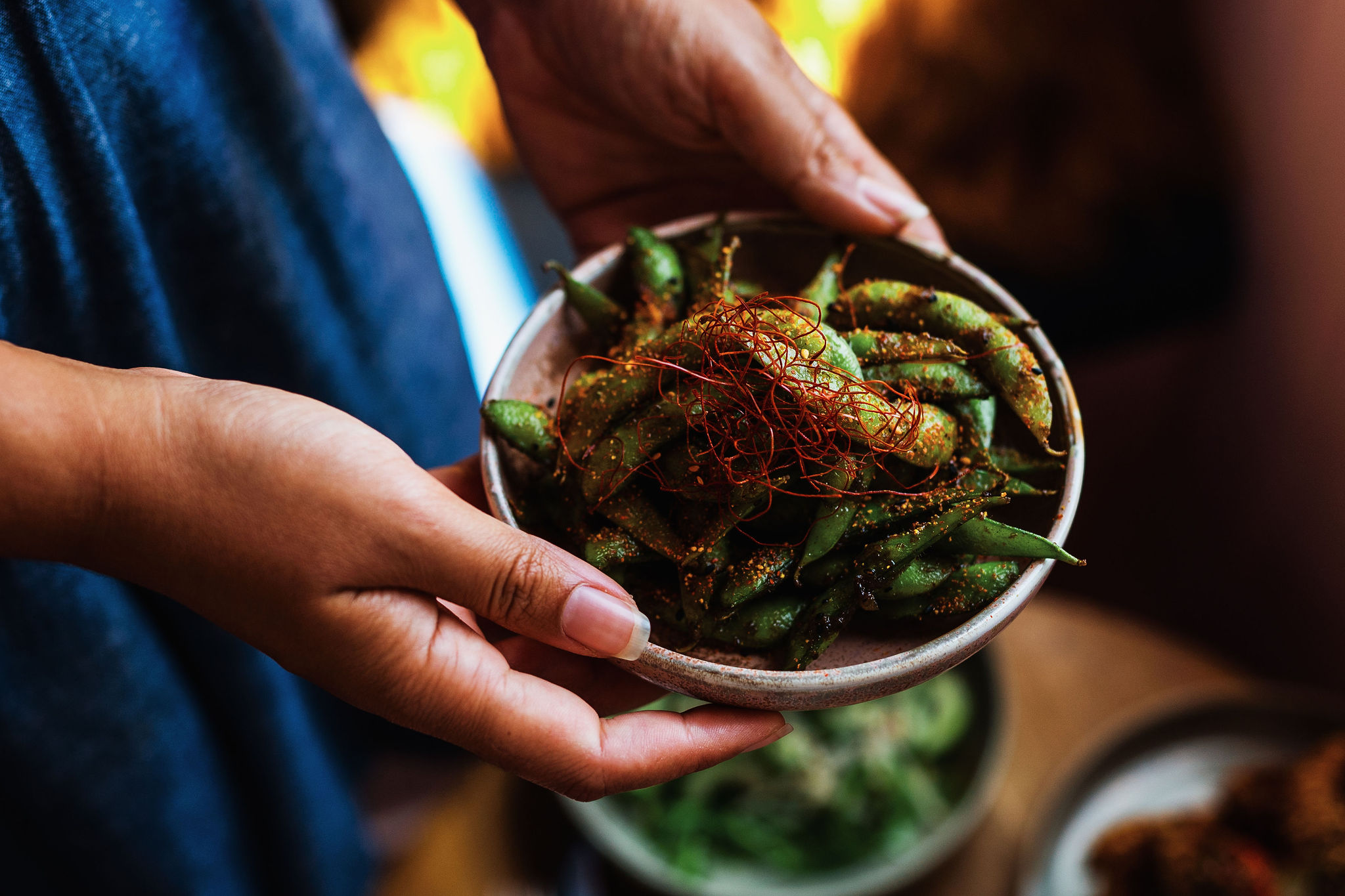Hands hold a small white bowl of seasoned green beans topped with red seasoning.