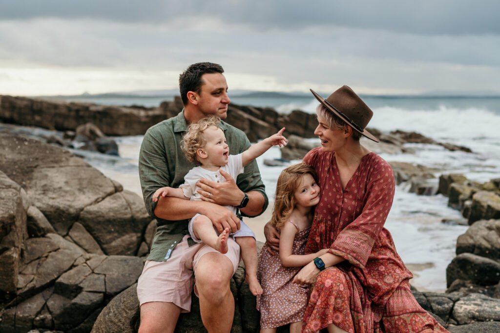 A family of four sits on a rocky shoreline by the ocean, with the father holding a baby and the mother wearing a hat beside their young daughter.