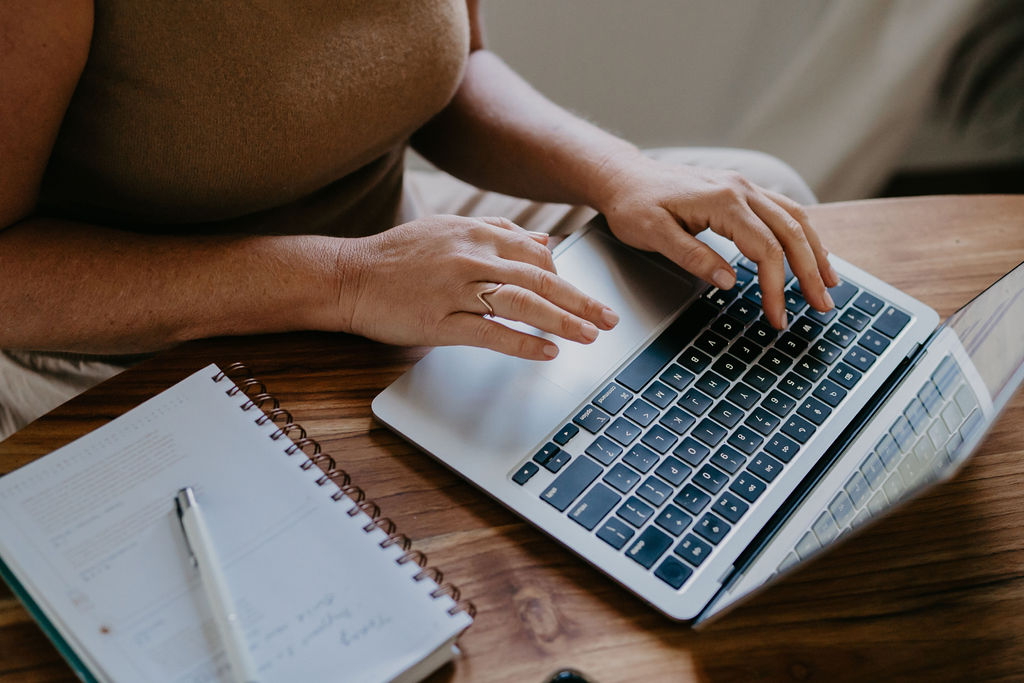 Hands typing on a laptop keyboard beside a notebook and pen on a wooden desk.