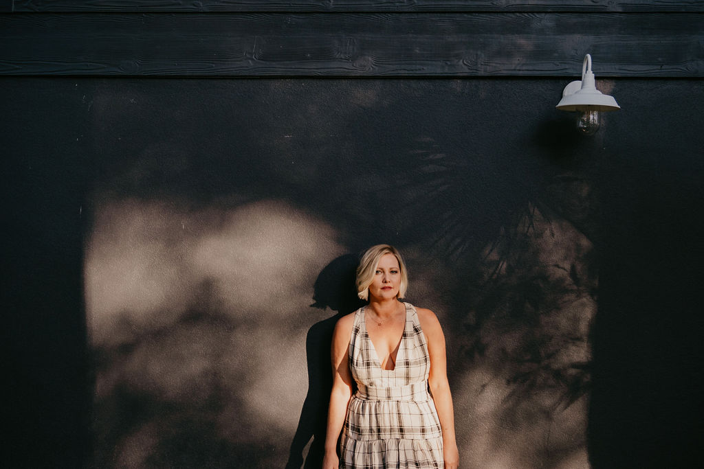 A blonde woman wearing a checkered dress stands against a dark wall with dappled sunlight and a wall-mounted light above.