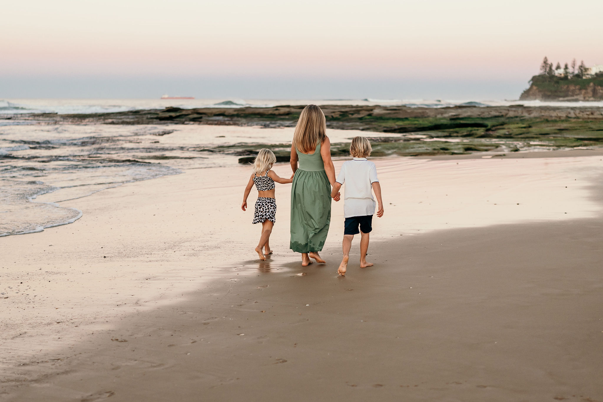 A woman in a green dress walks along a sandy beach with three children toward the water at sunset.