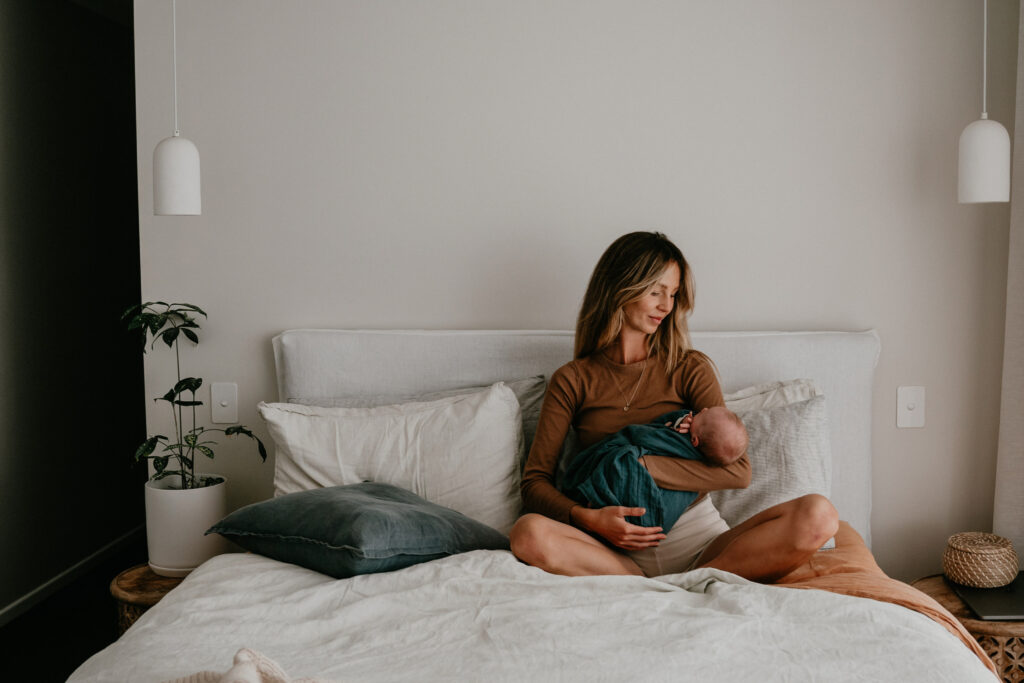 A woman sits on a bed cradling a baby wrapped in a teal blanket in a minimalist, cozy bedroom.