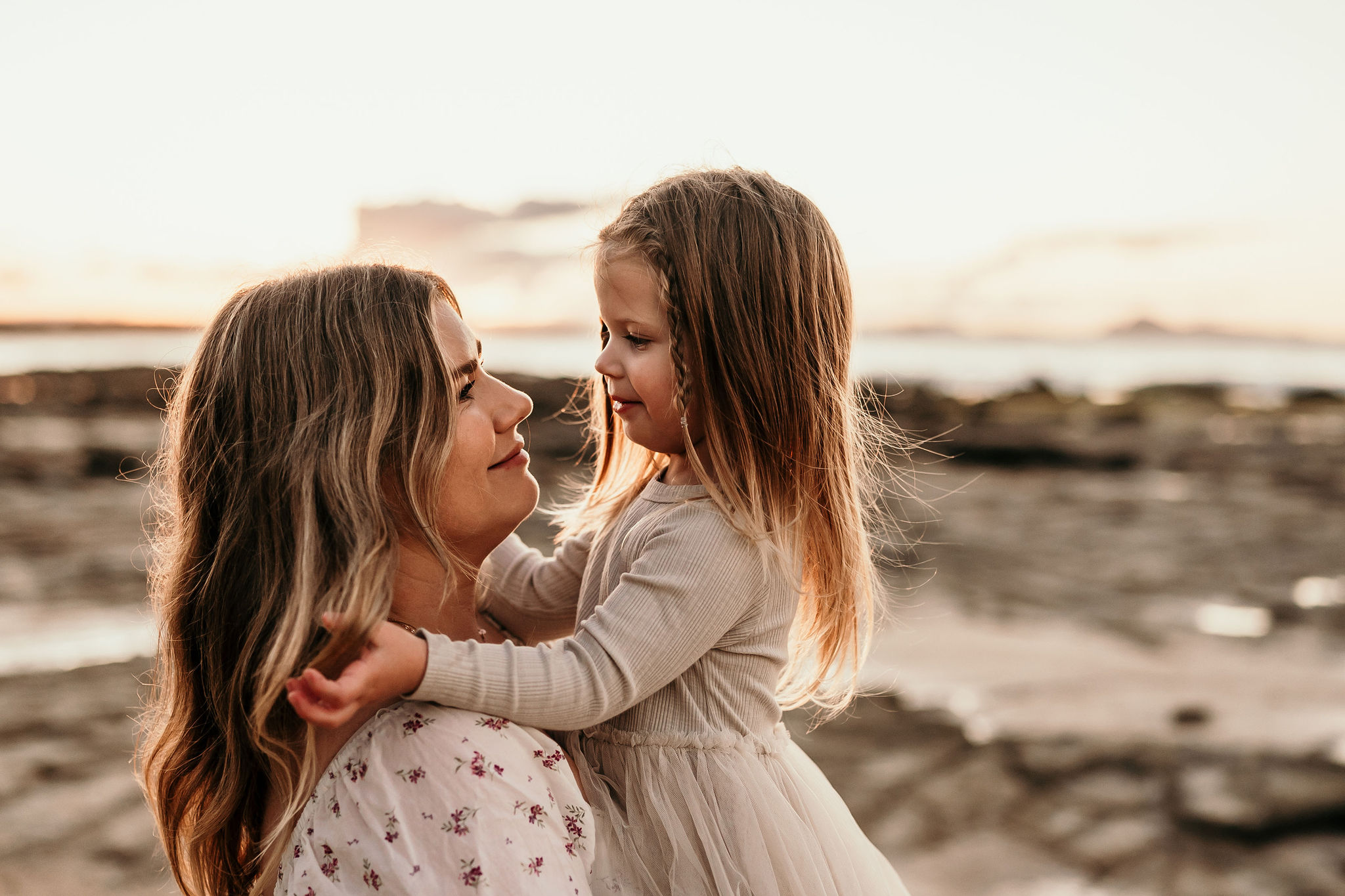 A mother and daughter share a hug on a rocky beach at sunset, smiling at each other.