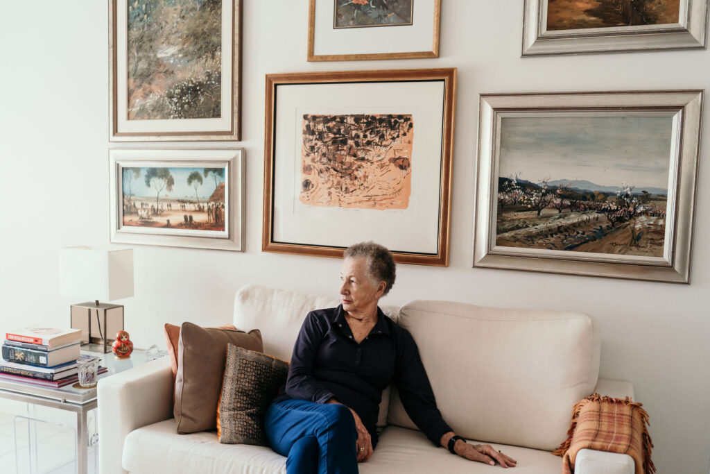 A man sits on a beige sofa in a living room with multiple framed landscape paintings on the wall behind him.