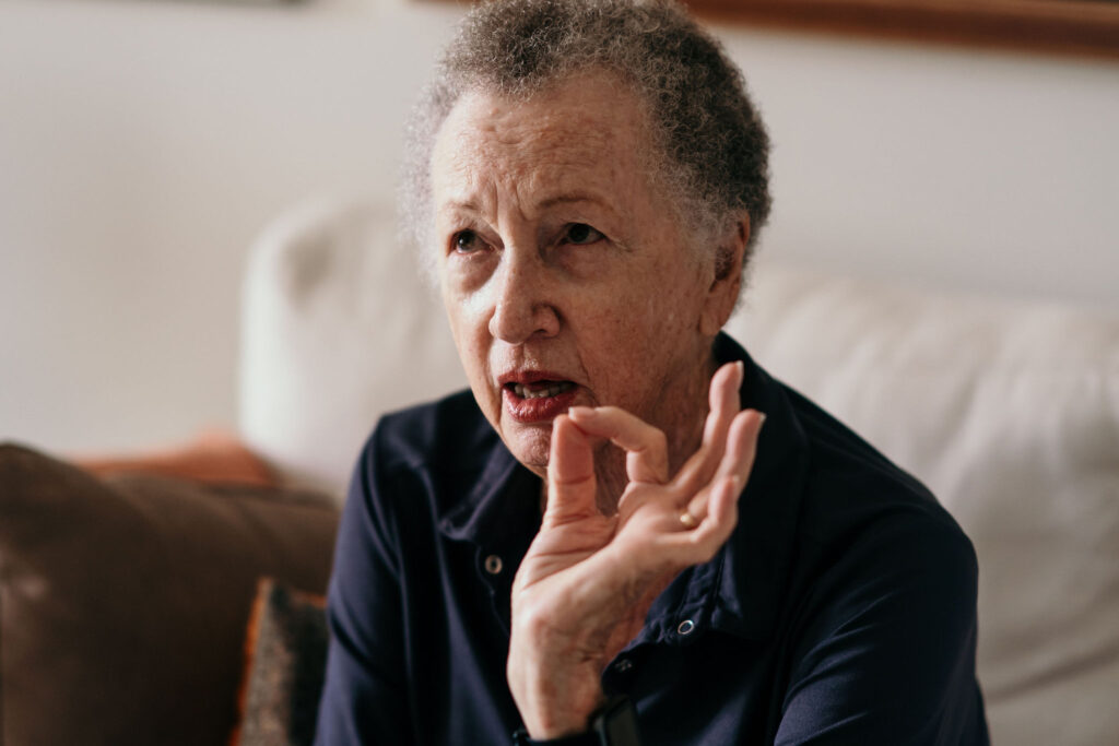An elderly man with gray hair sits on a couch in a living room, gesturing with his hand near his mouth.