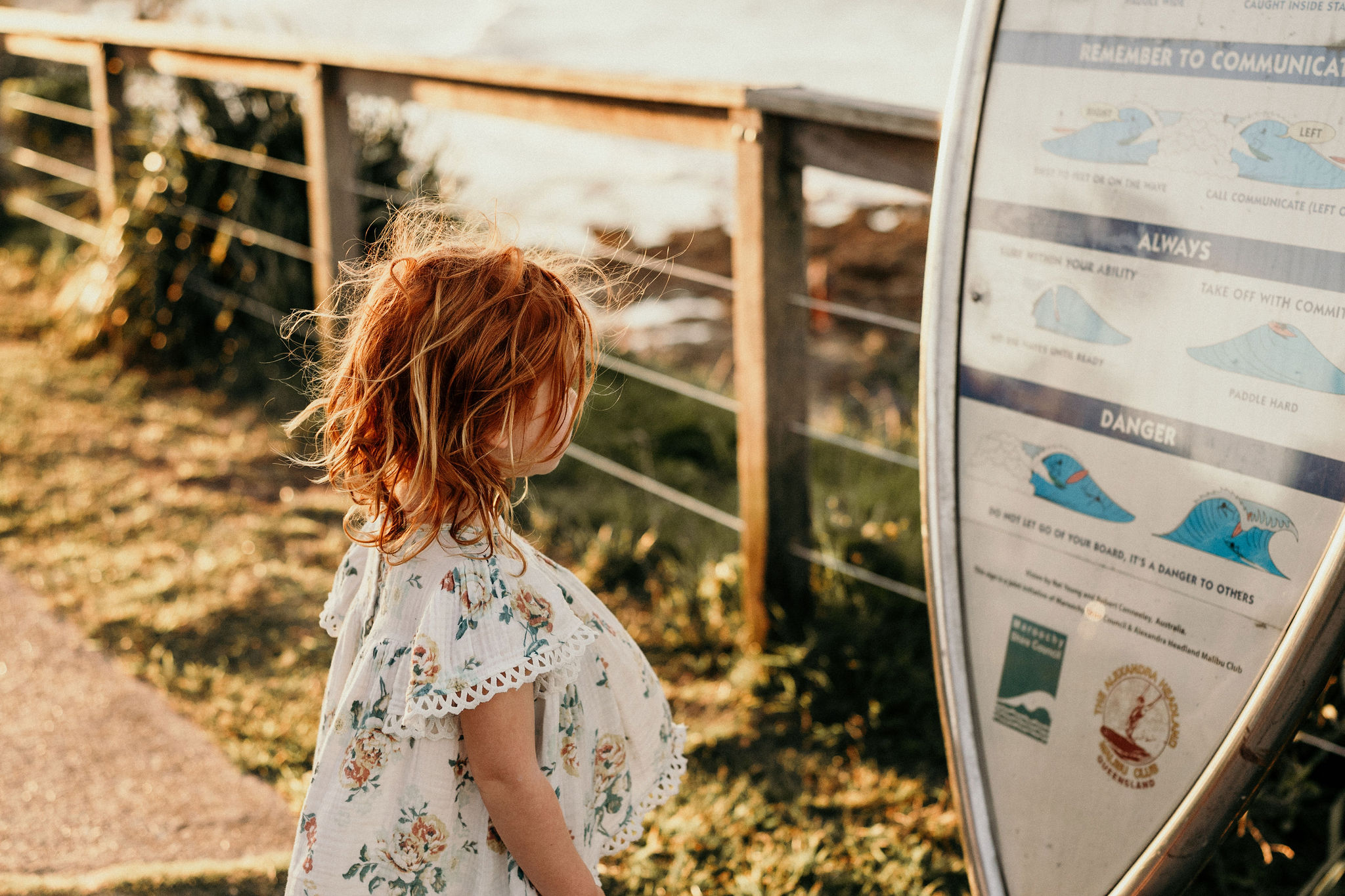 A young girl with red hair wearing a floral dress stands outdoors, looking at a tall information sign.
