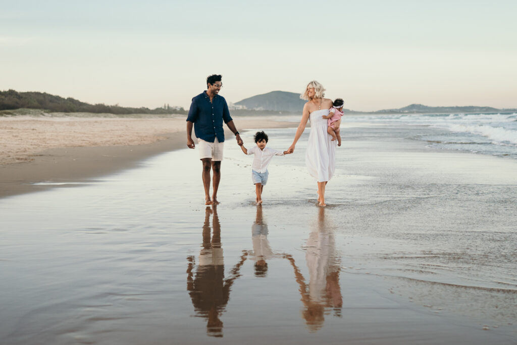 A family of four walks hand-in-hand along the shoreline, their reflections visible on the wet sand.