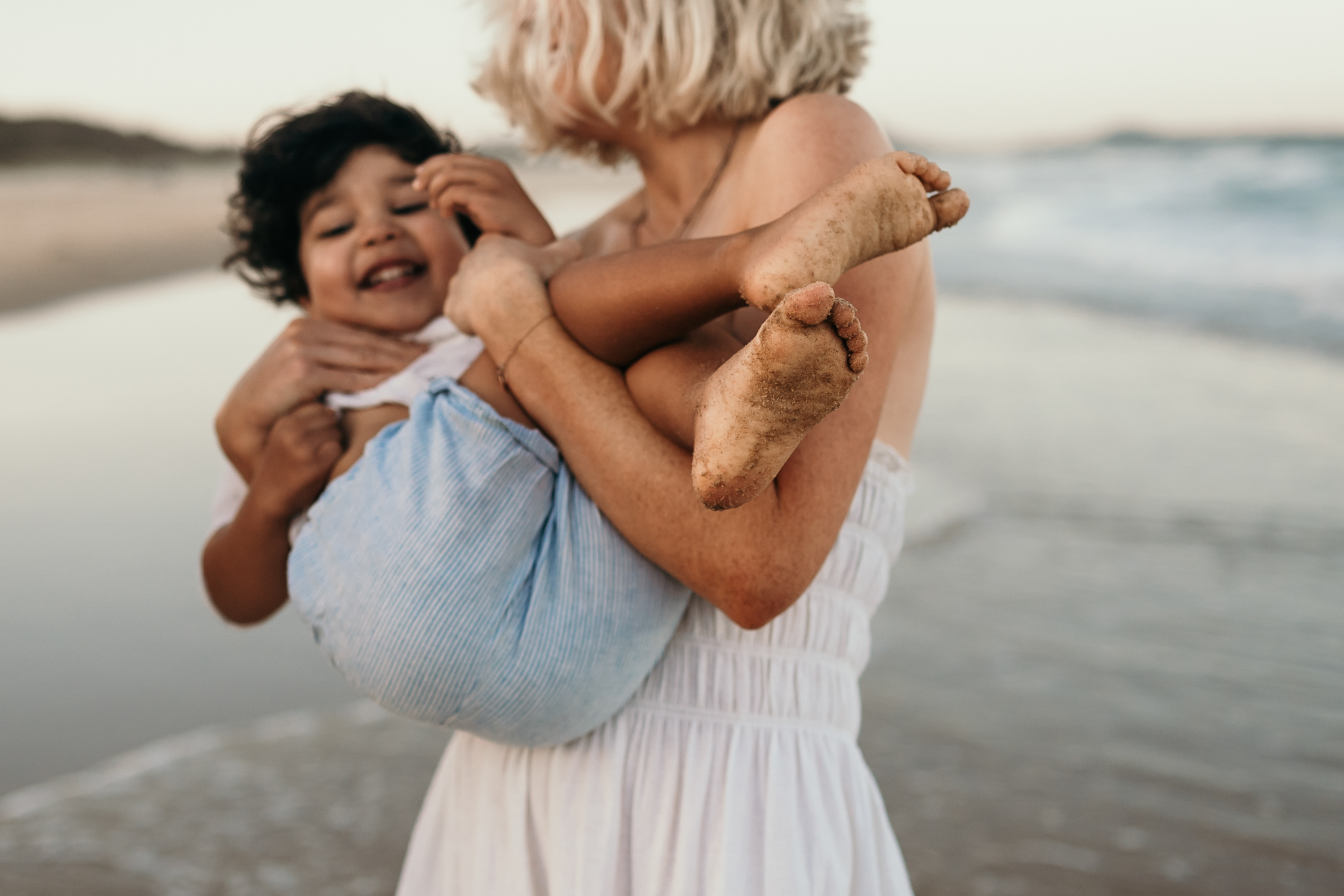 A woman in a white dress cradles a laughing baby at the beach.