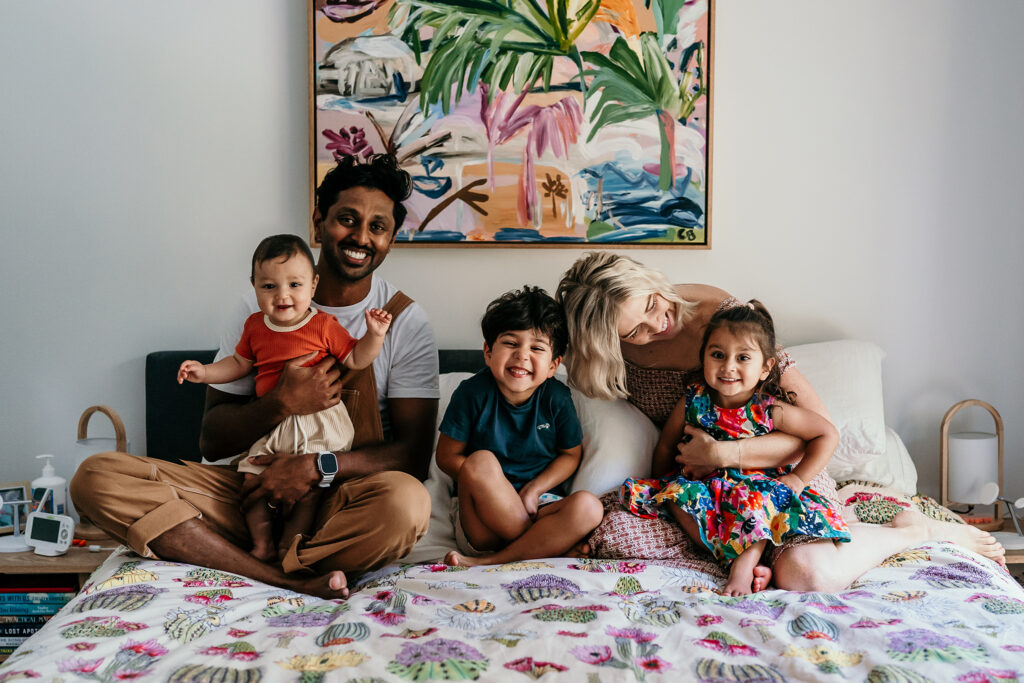 A family of five sits on a bed with a floral quilt, smiling at the camera.