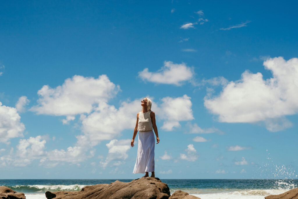 A person in a white dress stands on a rocky shore by the sea under a blue sky with white clouds.