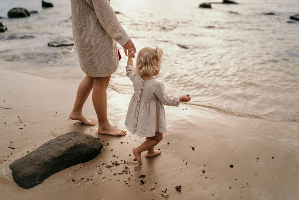 A barefoot adult and a small child walk along a wet sandy beach at the water's edge, holding hands.