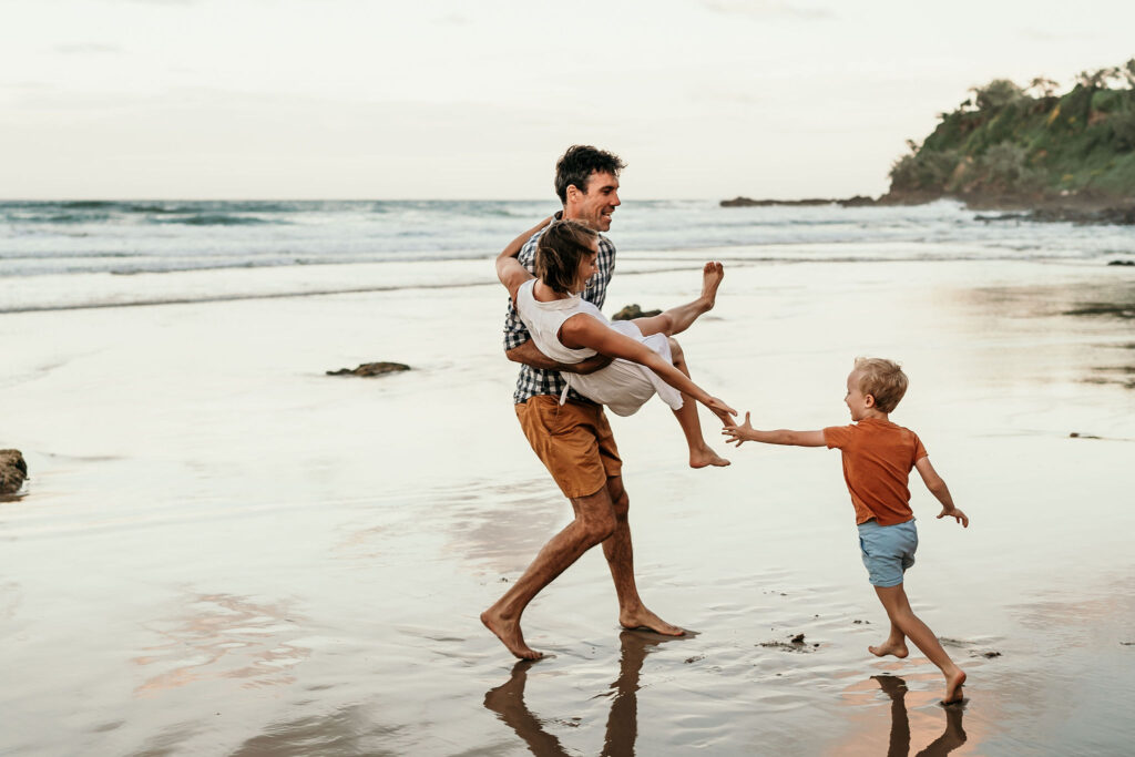 A man in a white shirt and brown shorts carries a young child on a sandy beach as another child in an orange shirt and blue shorts runs toward them along the wet shoreline.