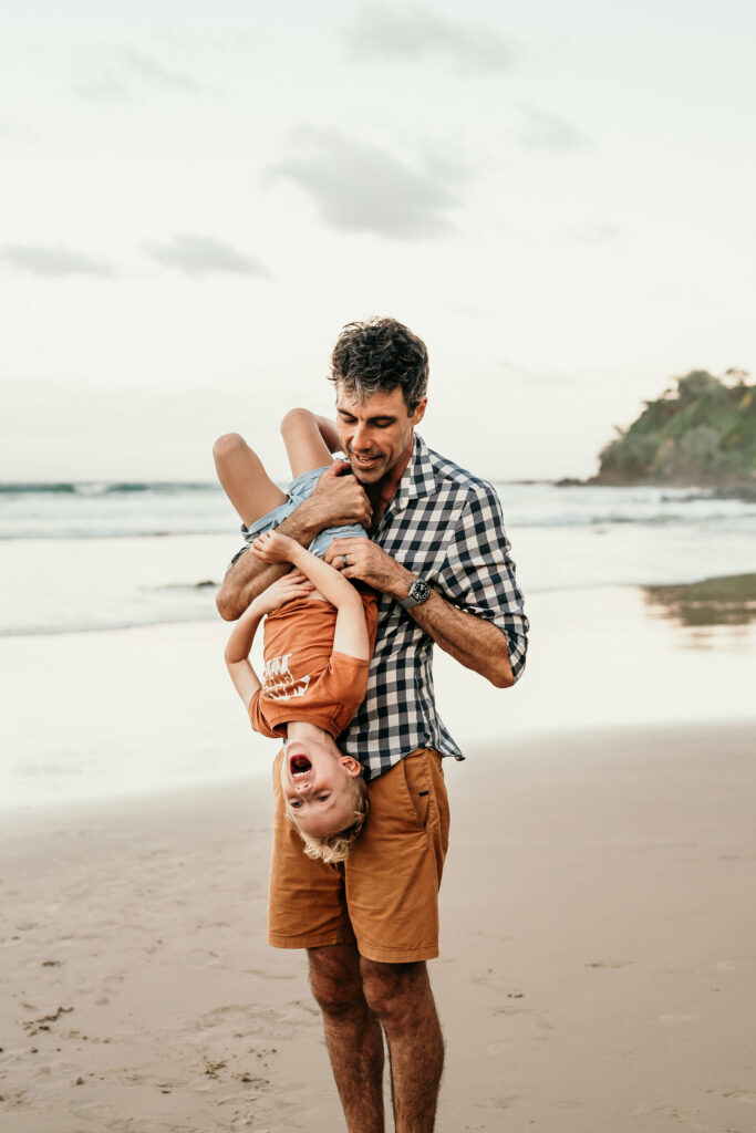 Man on a sandy beach holding a baby upside down in a playful pose with the ocean and coastline in the background.