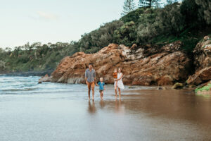 Three people walk along a wet sandy beach with rugged rocky cliffs and trees in the background.
