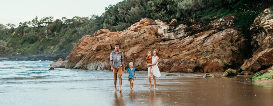 Three people walk along a wet sandy beach with rugged rocky cliffs and trees in the background.