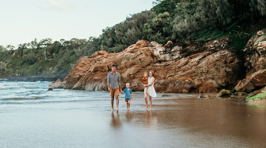Three people walk along a wet sandy beach with rugged rocky cliffs and trees in the background.