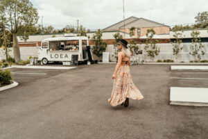A woman in a flowing floral dress walks away from a white bus in a parking lot.
