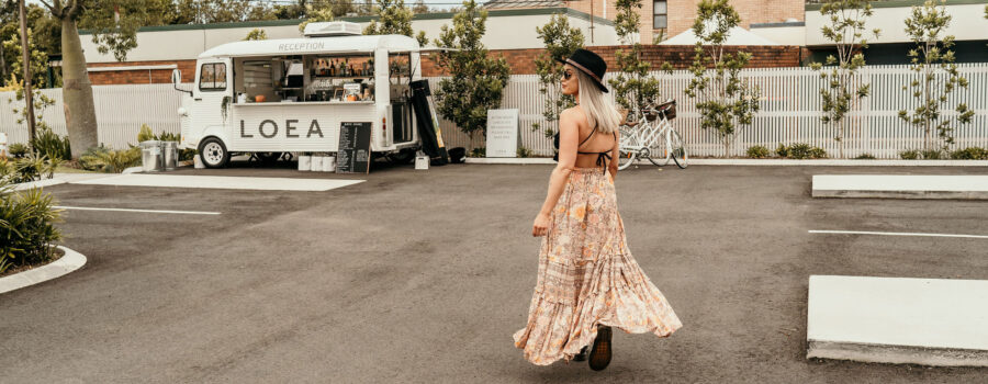 A woman in a flowing floral dress walks away from a white bus in a parking lot.