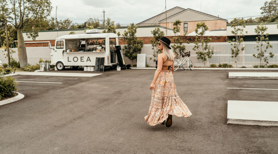 A woman in a flowing floral dress walks away from a white bus in a parking lot.