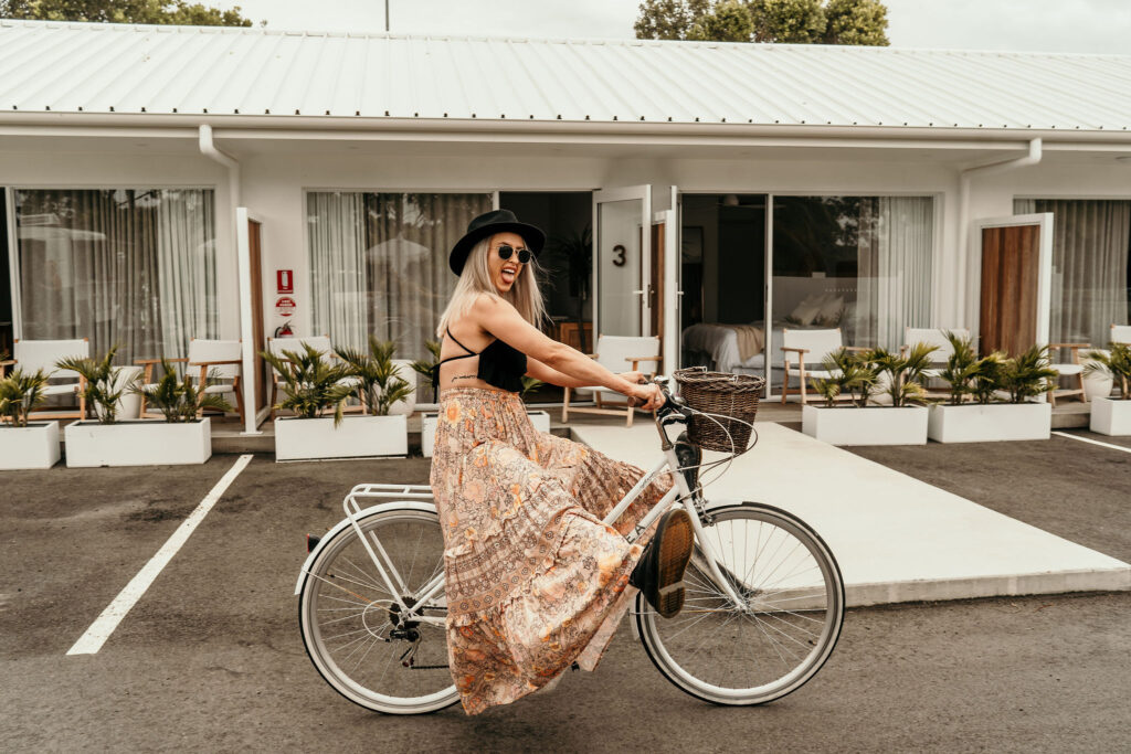 Woman wearing a long floral dress and sunglasses sits on a white bicycle in a parking lot in front of a row of storefronts.