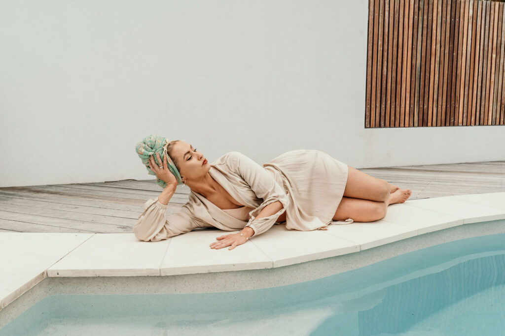 A woman in a beige robe lies on her side on a white poolside deck with a towel wrapped around her head.
