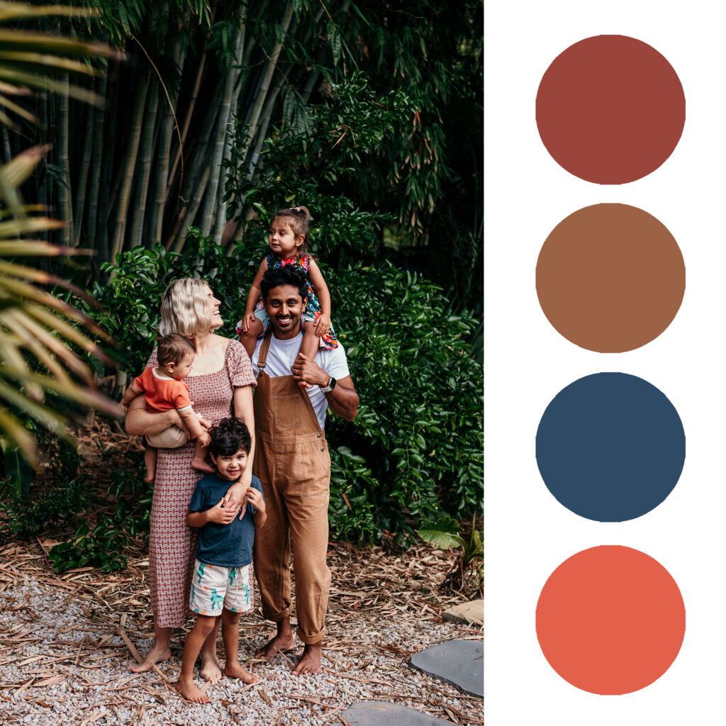 Family of four posing outdoors among trees, with a father in brown overalls carrying a child on his shoulders and a mother holding another child.